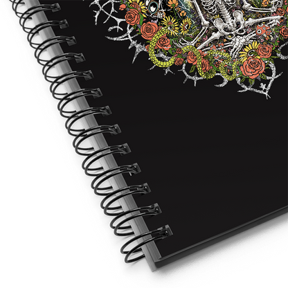 Detail of a spiral notebook journal with vibrant digital artwork of a meditating skeleton in a yoga pose amid colorful flowers and insects. A bluebird sits on the skeleton's shoulder. It's framed by a heart-shaped iron fence with snakes, roses, and gothic 'Manifest' text. Expressive eyes and a third eye symbolize spiritual awakening. Despite the edgy look, it carries an uplifting message of manifestation and abundance. © Donny Meloche, 2023.